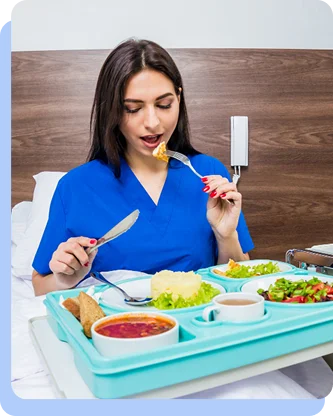 A woman in blue medical scrubs sitting on a hospital bed and eating a balanced meal from a blue plastic tray, representing structured nutrition in a healthcare environment.