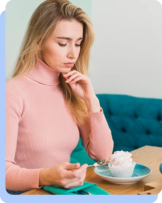 A woman in a pink turtleneck sitting at a table, looking down thoughtfully or hesitantly at a single cupcake on a blue plate.