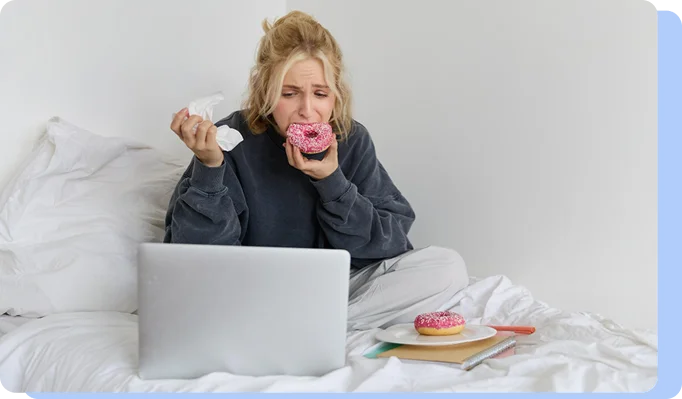 A distressed-looking woman sitting on a bed with a laptop, holding a crumpled tissue and crying while eating a pink frosted donut, depicting stress-related or emotional eating.