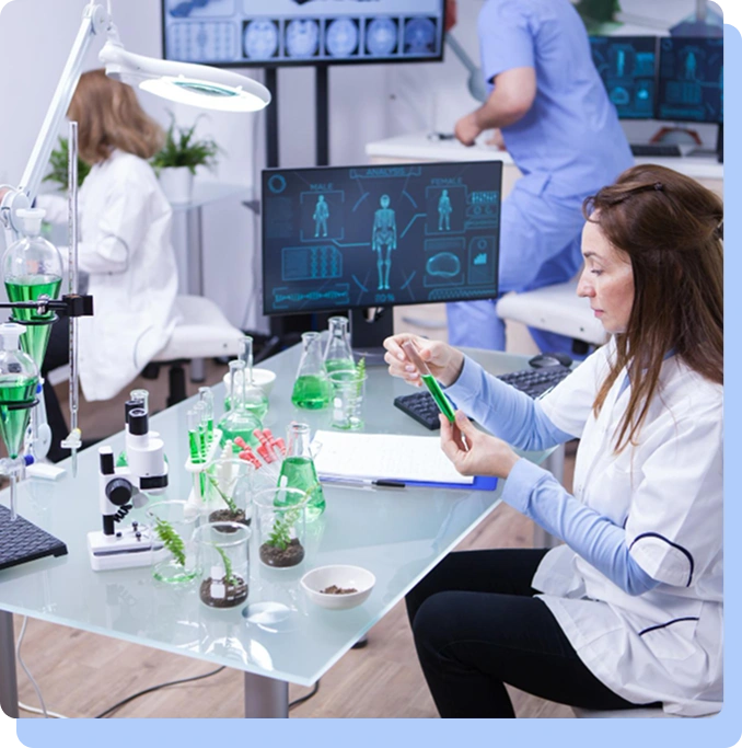 A wide shot of a high-tech laboratory where a female scientist in a white lab coat is seated at a glass table, inspecting a test tube filled with green liquid. In the background, another researcher in blue scrubs works near a large monitor displaying a digital human skeleton and biological data.