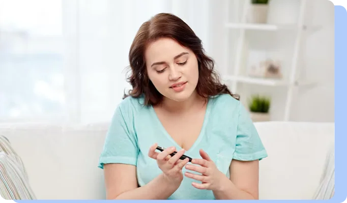 A woman in a light green shirt sitting on a couch and looking down at a hand-held glucose monitor while testing her blood sugar.