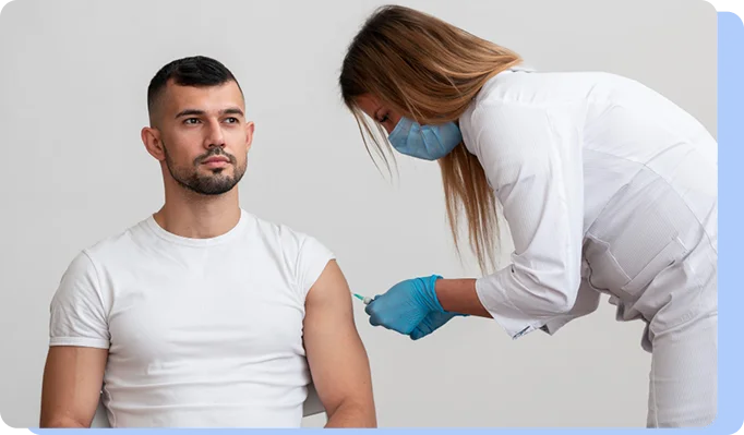 A healthcare professional in a mask and blue gloves administering an injection into the upper arm of a man in a white t-shirt.