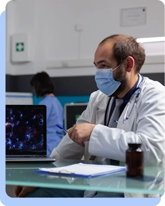 A doctor wearing a blue surgical mask and a white coat sits at a desk in a modern medical office, looking at a computer screen.