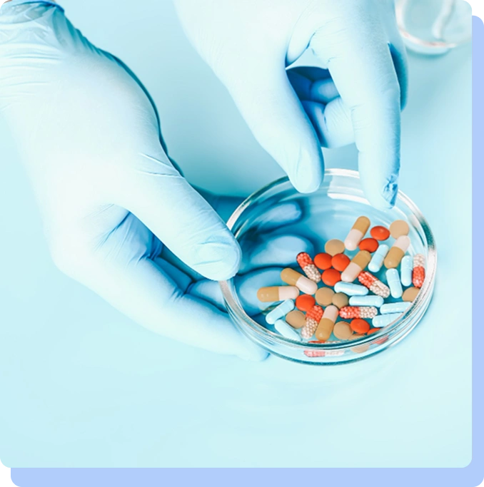 A top-down shot of a person wearing light blue medical gloves, carefully picking through a glass petri dish filled with a variety of colorful capsules and pills.