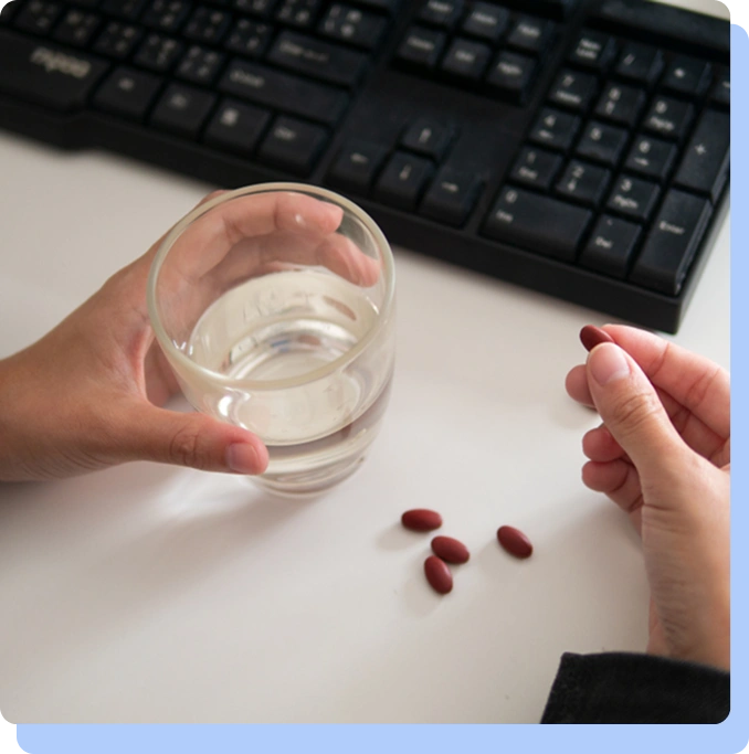 An overhead shot of a person’s hands at a white desk next to a computer keyboard. One hand holds a glass of water, and the other holds a single red pill, with several other red pills scattered on the desk surface.