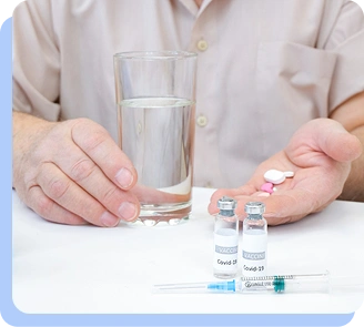A close-up of a person's hands on a table next to a glass of water, two small medical vials, and a few loose pills.