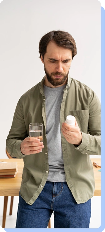 A man in a green button-down shirt standing in a kitchen, looking down thoughtfully at a glass of water and a small white pill bottle he is holding.