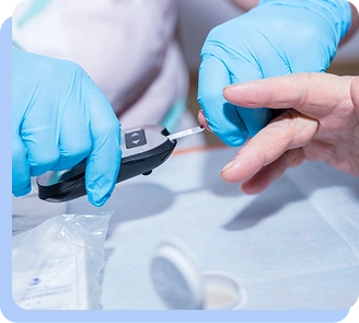 A healthcare provider in blue gloves using a portable glucose meter (glucometer) to test a patient's blood sugar level from a finger prick.