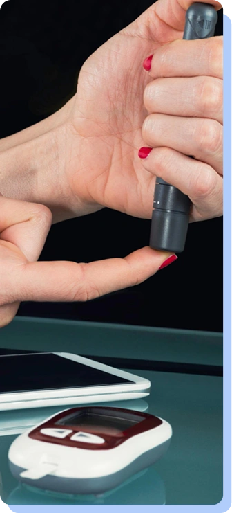 A person’s hands are shown using a lancing device on a fingertip. In the background, a digital tablet and a glucose meter sit on a reflective green surface.