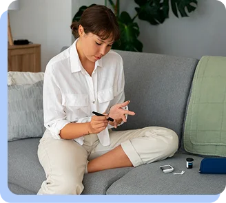 A woman sitting on a grey sofa in a brightly lit room, carefully using a lancing device on her finger.