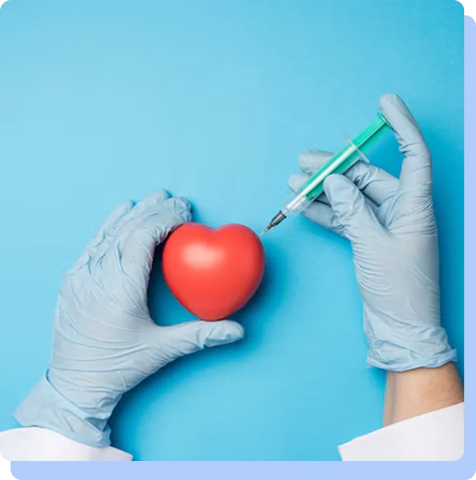 A top-down view of a pair of hands in blue medical gloves on a light blue surface. One hand holds a small red heart model while the other hand points a medical syringe toward it.