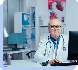 A senior doctor with glasses and a stethoscope sitting at a desk in a medical office, looking toward the camera.