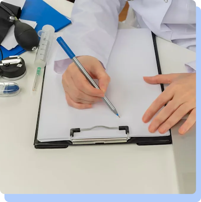 An overhead view of a medical professional in a white lab coat sitting at a desk and writing on a white piece of paper attached to a black clipboard. A blood pressure cuff and a syringe are visible on the desk nearby.