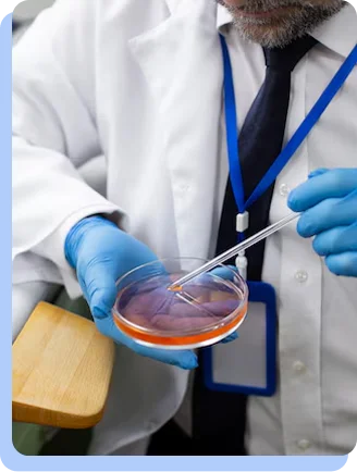 A close-up of a scientist in a white lab coat and blue gloves using a pipette to drop liquid into a Petri dish.