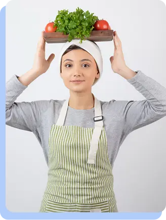 A woman in a grey long-sleeved shirt and a green-and-white striped apron, balancing a wooden cutting board with fresh green herbs on top of her head.