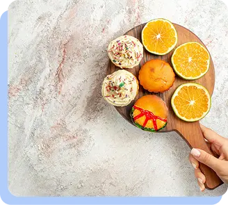 A close-up, top-down view of a hand holding a wooden paddle featuring several decorated cupcakes and sliced oranges.
