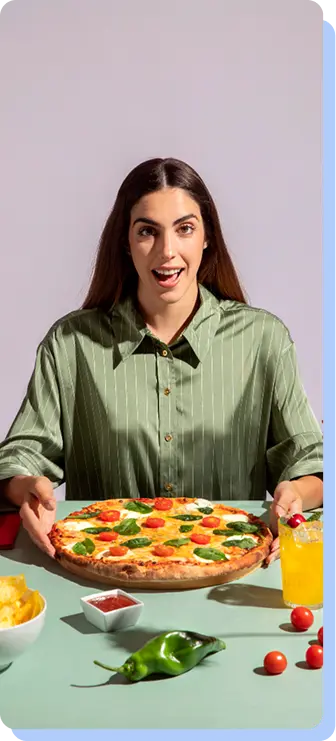 A woman in a green button-down shirt sitting at a table, looking excited with an open mouth as she holds a large pizza topped with tomatoes and basil.