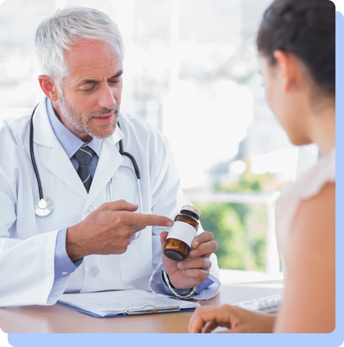 A senior male doctor with grey hair and a white lab coat sitting at a desk, pointing to the label on a brown medicine bottle while explaining its contents to a female patient whose back is to the camera.