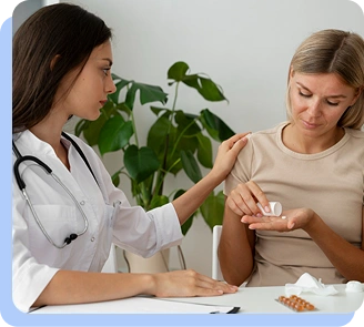A female doctor in a white coat sitting at a desk, comfortingly placing a hand on the shoulder of a patient who is holding a small medication bottle.