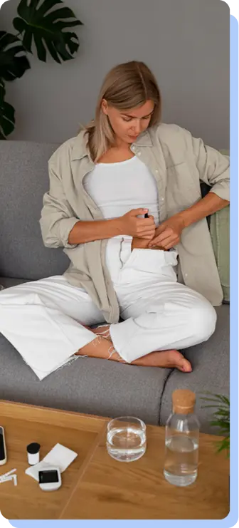 A woman in a beige shirt and white pants sitting on a grey sofa, holding a medication pen to her abdomen to administer an injection. A glass of water and a bottle are on the table in front of her.