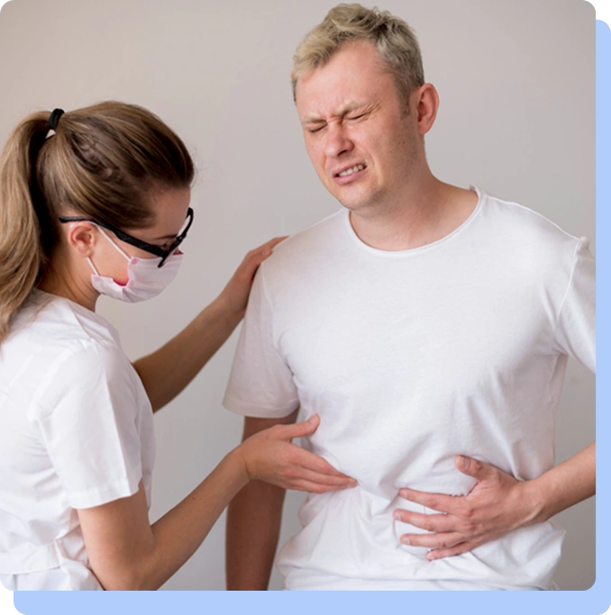 A woman in medical attire and a face mask providing care to a man in a white t-shirt. The man has a pained facial expression and is clutching his abdomen, while the medical worker places a supportive hand on his shoulder and examines his side.
