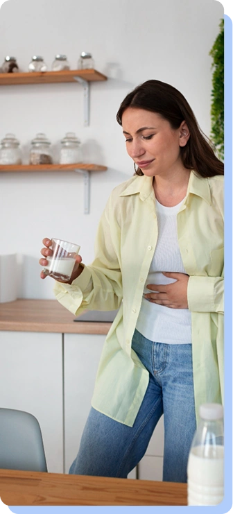 A woman in a pale yellow shirt standing in a kitchen, looking down with a pained expression while holding a glass of water and clutching her stomach.
