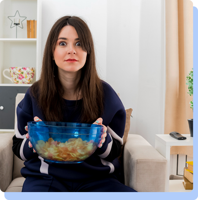 A woman sitting on a couch, holding a large blue glass bowl filled with chips or snacks with a neutral, slightly wide-eyed expression, illustrating the concept of fullness and snacking habits.