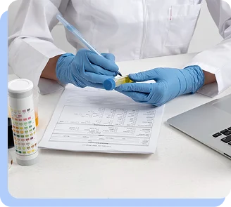 A close-up of a researcher wearing blue surgical gloves, using a pen to record data on a clinical form next to a laptop.
