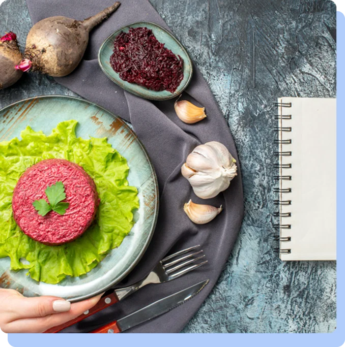 A top-down culinary shot of a healthy beet tartare dish served on a bed of lettuce. The scene is styled on a textured blue surface with raw ingredients like whole beets, garlic cloves, a side dish of beet salad, and a blank spiral notebook with a fork and knife.