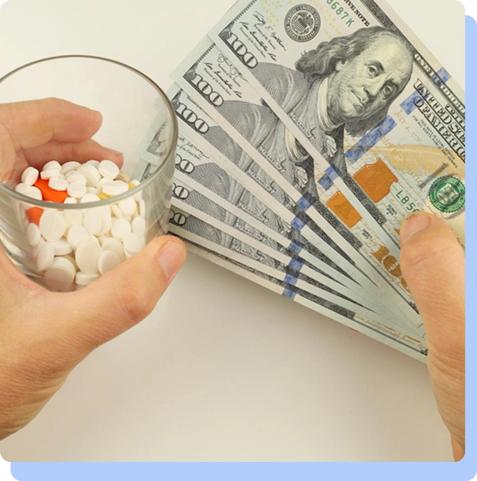 A close-up of a person’s hands holding a glass filled with white and orange pills next to a fan of $100 bills.