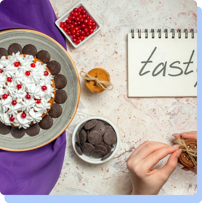 A flat-lay photograph of various sweets on a textured surface, featuring a cake with white frosting and red berries, a bowl of chocolate discs, red currants, and a person's hands untying a small gift-wrapped cookie. A notebook next to the food has the word "tasty" written on it.