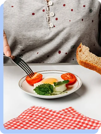 A close-up of a person about to eat a healthy plate containing a fried egg, sliced tomatoes, and greens, with a piece of whole-grain bread held nearby.