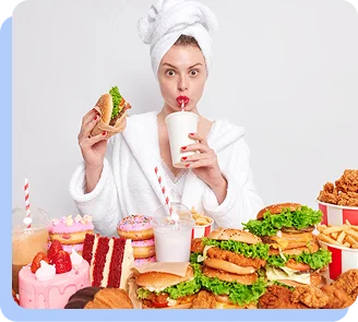 A woman in a white bathrobe sitting at a table overflowing with junk food, including multiple burgers, fries, cakes, and donuts, while she eats a burger and drinks from a large cup.