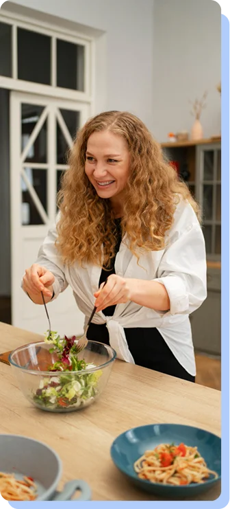 A woman with long curly hair smiling as she tosses a fresh green salad in a large glass bowl in a bright kitchen.