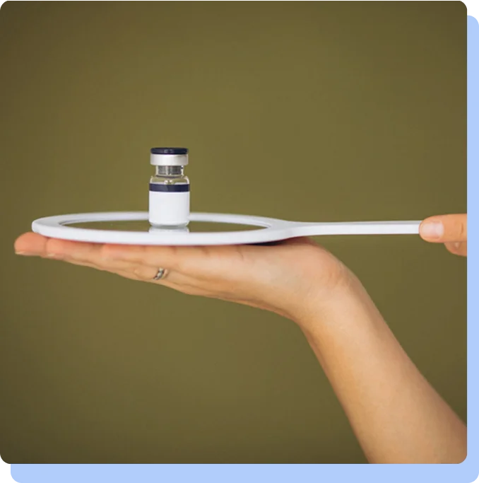 A hand holding a small white hand-mirror horizontally, with a single small medical vial balanced on the reflective surface.