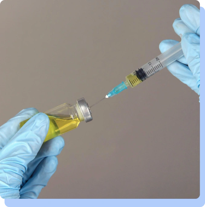 A close-up of hands in blue medical gloves using a syringe to draw yellow liquid from a small glass vial.