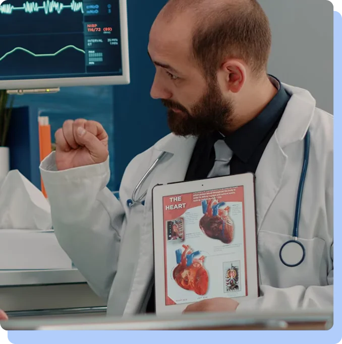 A male doctor with a beard wearing a white lab coat and stethoscope, holding up a tablet that displays detailed medical diagrams of the human heart and circulatory system while gesturing with his other hand.