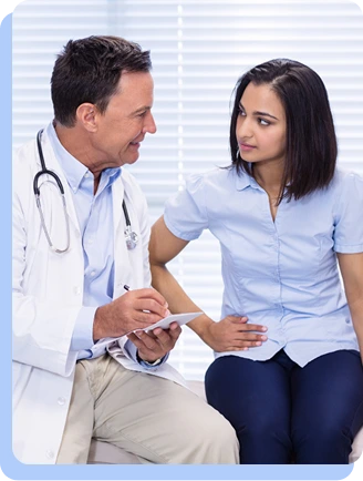 A male doctor in a white coat sitting and talking with a female patient in a light blue shirt; they appear to be in a serious but calm medical discussion.