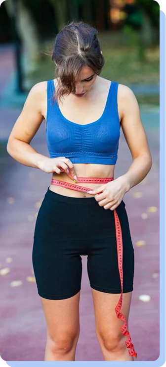 A woman in athletic wear (a blue sports bra and black shorts) standing outdoors, using a pink measuring tape to check her waist circumference.