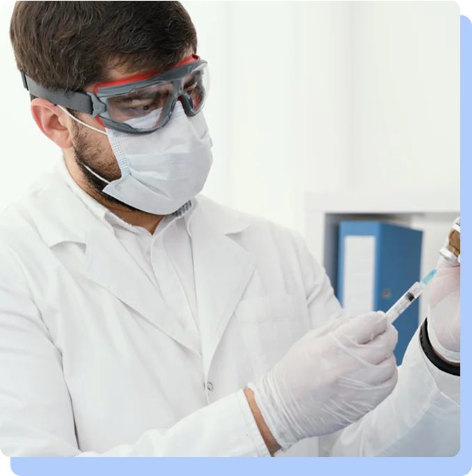 A close-up of a medical professional wearing a face mask, safety goggles, and white gloves, carefully drawing liquid from a glass vial into a syringe.