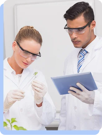 A man and a woman in lab coats working together; the woman holds a small plant seedling while the man holds a digital tablet.