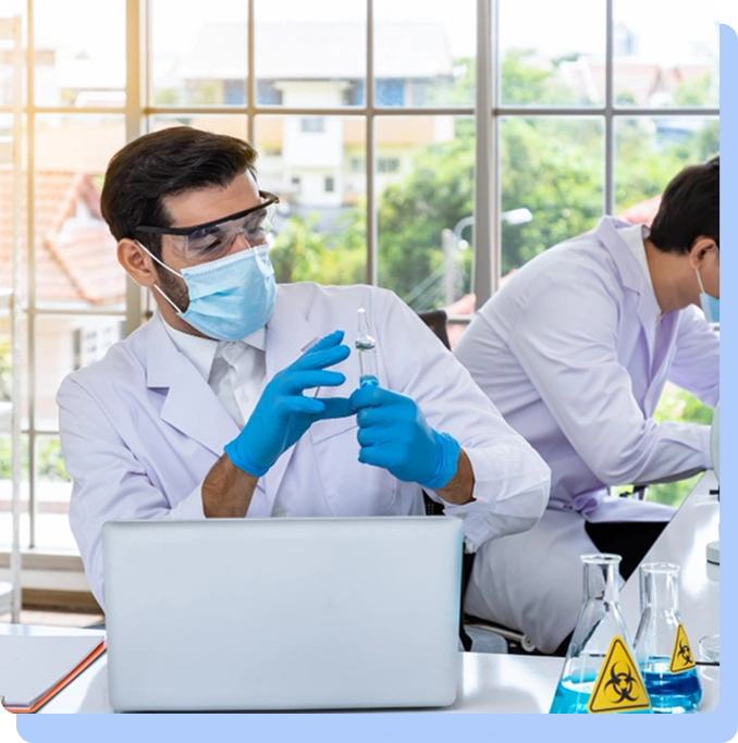 Two scientists in a bright, modern laboratory with large windows. The scientist in the foreground wears a face mask, safety glasses, and blue gloves while examining a liquid sample in a vial. A laptop and various beakers labeled with biohazard symbols are on the desk.