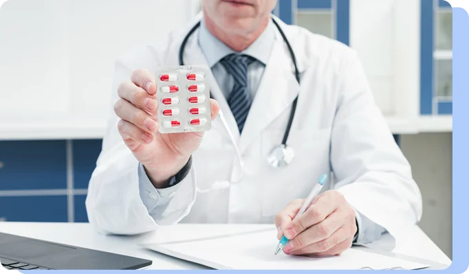 A doctor in a lab coat sitting at a desk, holding a blister pack of red and white pills in one hand and a pen in the other.