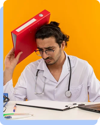 A male doctor in a lab coat looking stressed or overwhelmed, holding a red binder against his head while sitting at a desk with a stethoscope around his neck.