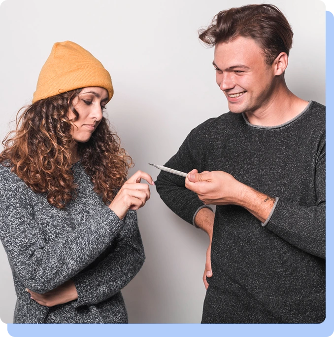 A man smiling while showing an injection pen to a woman wearing an orange beanie.