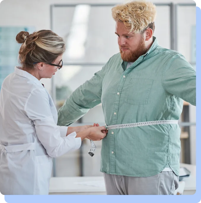 A female doctor in a white lab coat using a measuring tape to measure the waistline of a male patient in a green button-down shirt.