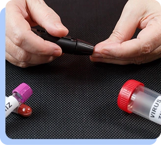 A close-up of a person's hands holding a small medical device, with blood collection tubes visible in the foreground.
