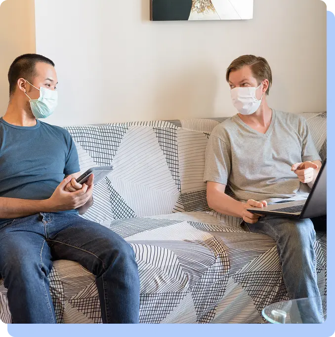 Two men wearing protective face masks sitting on a couch and looking at a digital tablet and laptop, illustrating the accessibility of medical information and telehealth.