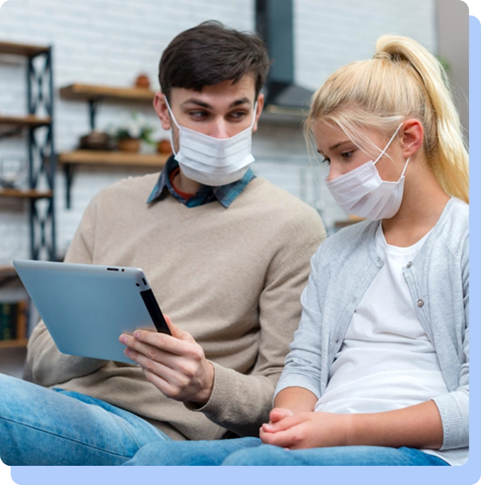 A man and woman wearing protective face masks looking at a digital tablet together in a home-like setting.
