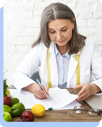 A woman with grey hair sitting at a wooden table, writing on a meal planning sheet. Fresh vegetables, including peppers and a lemon, are spread out on the table around her.